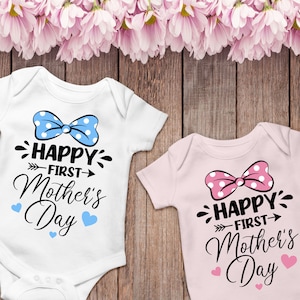 Peut inclure: Deux bodies blancs pour b&eacute;b&eacute; avec un n&oelig;ud et le texte "Happy First Mother's Day". Un body a un n&oelig;ud bleu et des c&oelig;urs bleus, l'autre a un n&oelig;ud rose et des c&oelig;urs roses.