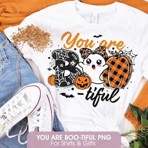 Può includere: Una t-shirt bianca con un design grafico che riporta il testo You are BOO-tiful. Il disegno include una lettera B nera con ragnatele, un piccolo fantasma con un fiocco leopardato e una zucca a quadri arancioni e neri con piccoli pipistrelli.