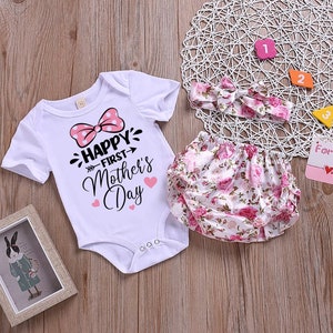 Peut inclure: Un body blanc pour b&eacute;b&eacute; avec un n&oelig;ud rose &agrave; pois et le texte "Happy First Mother's Day". Un bandeau blanc &agrave; imprim&eacute; floral rose et une couche blanche &agrave; imprim&eacute; floral rose.