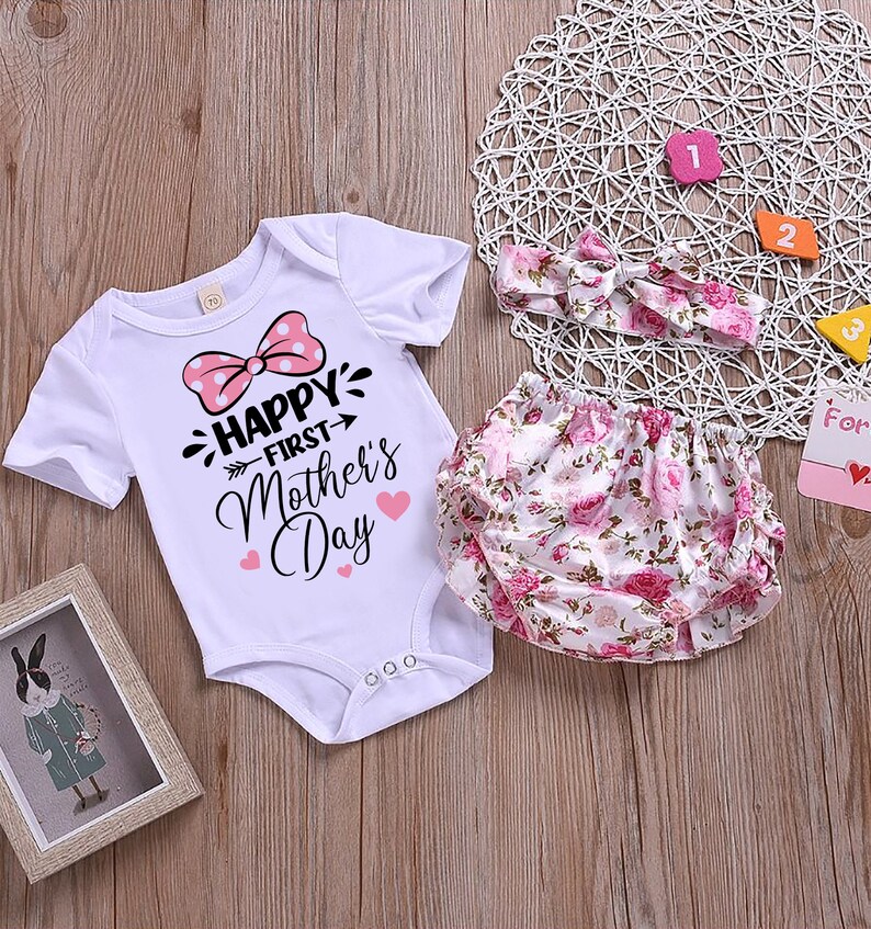 Peut inclure: Un body blanc pour b&eacute;b&eacute; avec un n&oelig;ud rose &agrave; pois et le texte "Happy First Mother's Day". Un bandeau blanc &agrave; imprim&eacute; floral rose et une couche blanche &agrave; imprim&eacute; floral rose.