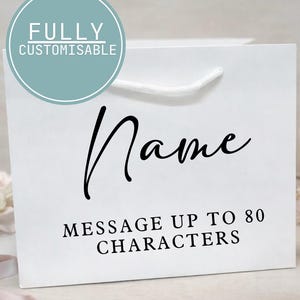 May include: White gift bag with a white rope handle. The bag features the word "Name" in a black script font, and the text "MESSAGE UP TO 80 CHARACTERS" in a black sans-serif font. A teal circle with the words "FULLY CUSTOMISABLE" is in the upper left corner.