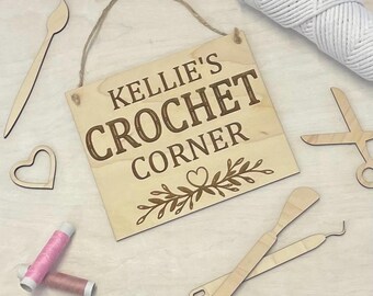 May include: A wooden sign with the text "Kellie's Crochet Corner" in brown lettering. The sign is decorated with a small heart and leafy vines. The sign is surrounded by various crochet supplies, including yarn, needles, and scissors.