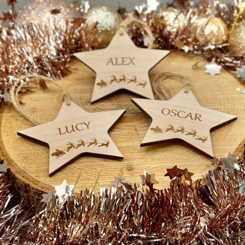 May include: Three wooden star-shaped ornaments with names "Alex", "Lucy", and "Oscar" engraved on them. Each star features a small engraved image of Santa Claus in a sleigh pulled by reindeer.
