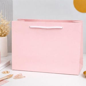 May include: A pale pink gift bag with a white handle rests on a white surface. A lit pink candle, a pen, and a small gold dish with pink sweets are also on the surface. A small vase with dried flowers is in the background.