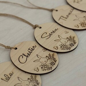 May include: Four wooden Easter egg ornaments with bunny and floral designs. Each ornament has a different name written on it: Isla, Chester, Seren, and Thomas.