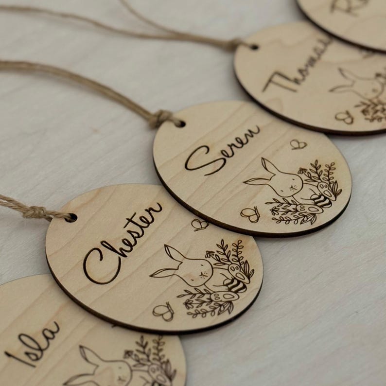 May include: Four wooden Easter egg ornaments with bunny and floral designs. Each ornament has a different name written on it: Isla, Chester, Seren, and Thomas.