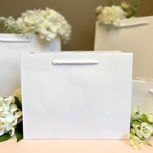 May include: A collection of white gift bags with white rope handles, arranged on a pink surface. White flowers are placed around the bags. The bags have a simple, elegant design.