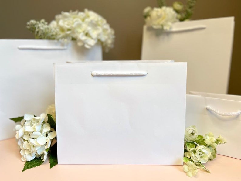 May include: A collection of white gift bags with white rope handles, arranged on a pink surface. White flowers are placed around the bags. The bags have a simple, elegant design.