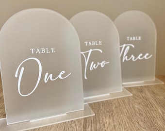 May include: Three frosted acrylic table numbers with arched tops. Each sign has the word "TABLE" at the top, with the numbers "One", "Two", and "Three" in a white script font. The signs are displayed on a wooden surface.