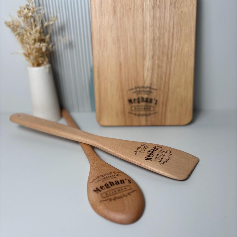 May include: A light brown wooden cutting board and two wooden kitchen utensils, a spoon and a spatula, each engraved with the text Meghan's Kitchen and Homemade with Love. The items are arranged together, with the utensils resting in front of the board.