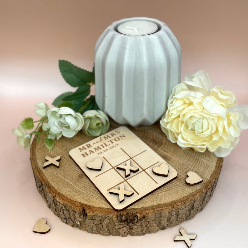 K&ouml;nnte beinhalten: Ein Tic-Tac-Toe-Spiel aus Holz mit dem Text "Mr. and Mrs. Hamilton 10.08.2024" oben. Das Spiel befindet sich auf einer Holzscheibe mit einem wei&szlig;en Kerzenhalter und wei&szlig;en Blumen.