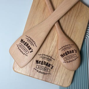 May include: A light-colored wooden cutting board, a flat wooden spatula, and a wooden spoon. Each item features a dark laser-engraved design with the text "Meghan's KITCHEN" and "HOMEMADE WITH LOVE" surrounded by decorative flourishes and lines.
