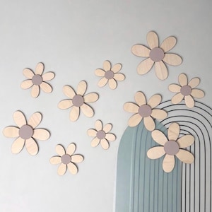 May include: Wooden daisy wall decorations in various sizes. Each flower has a light wood petal design with a muted purple centre. The daisies are arranged on a light grey wall, with a blue and black arched design in the background.