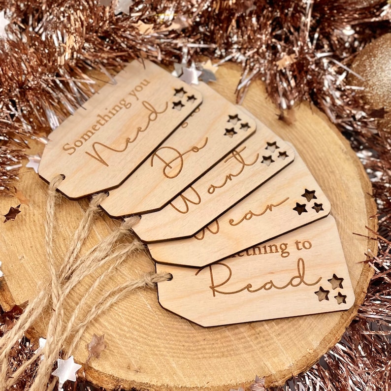 May include: Five wooden gift tags with twine attached. The tags are shaped like a teardrop and have the words "Something you Need", "Do", "Want", "Wear", and "Something to Read" written on them. Each tag has a star design cut out.