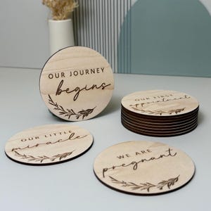 May include: A collection of wooden milestone discs with engraved text and botanical designs. The discs feature phrases such as "Our Journey Begins," "Our Little Miracle," "We Are Pregnant," and "Our First Appointment." The discs are light brown with dark brown edges.