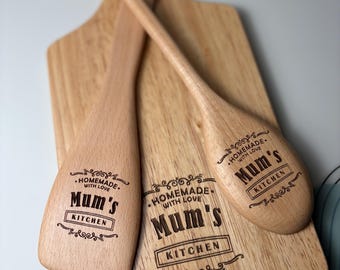 May include: Wooden kitchen utensils and cutting board. The cutting board and two utensils are engraved with the words "Mum's Kitchen" and "Homemade with Love" in a decorative font. The wood has a light brown color.