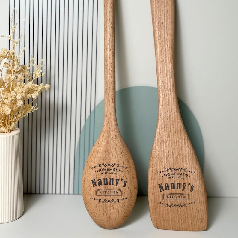 May include: Two light brown wooden kitchen utensils, a spoon and a spatula, each engraved with the text "Homemade with love Nanny's Kitchen" in a decorative black font. The utensils feature a smooth finish and natural wood grain patterns.