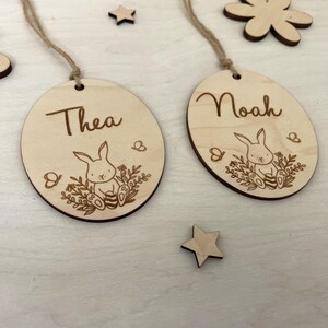 May include: Two wooden Easter egg ornaments with bunny and floral designs. The ornaments are personalized with the names "Thea" and "Noah".