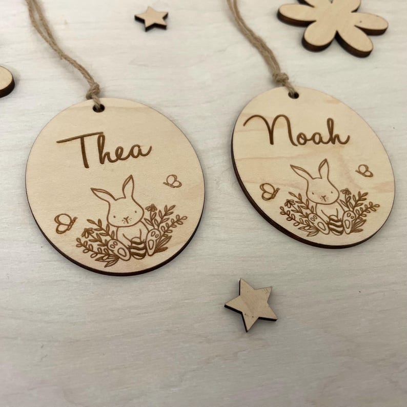 May include: Two wooden Easter egg ornaments with bunny and floral designs. The ornaments are personalized with the names "Thea" and "Noah".
