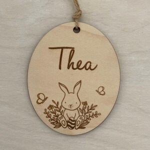 May include: A round wooden ornament with the name "Thea" engraved on it. A small bunny rabbit holding an egg is surrounded by flowers and butterflies.