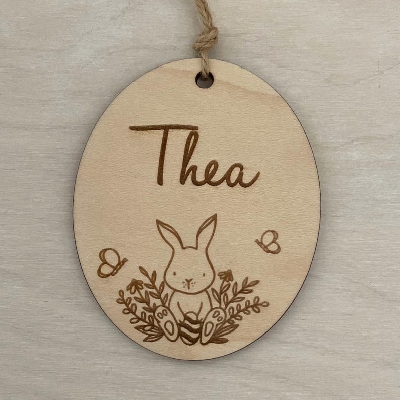 May include: A round wooden ornament with the name "Thea" engraved on it. A small bunny rabbit holding an egg is surrounded by flowers and butterflies.