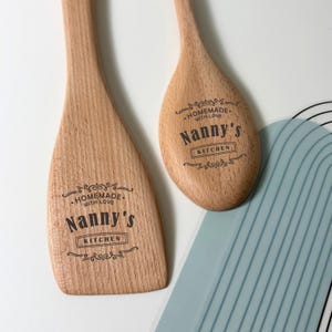 May include: Two light brown wooden kitchen utensils, a spatula and a spoon, each featuring a dark brown engraved design with the text "HOMEMADE WITH LOVE Nanny's KITCHEN" surrounded by decorative flourishes and lines on the flat surface.