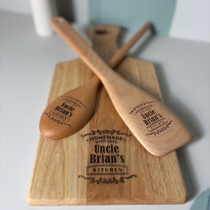 May include: A light-colored wooden cutting board and two wooden kitchen utensils, a spoon and a spatula, all engraved with the text "HOMEMADE WITH LOVE Uncle Brian's KITCHEN" and decorative flourishes. The items feature a smooth, natural wood grain.