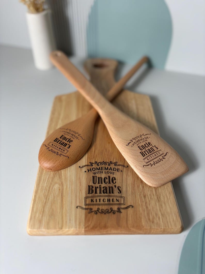 May include: A light-colored wooden cutting board and two wooden kitchen utensils, a spoon and a spatula, all engraved with the text "HOMEMADE WITH LOVE Uncle Brian's KITCHEN" and decorative flourishes. The items feature a smooth, natural wood grain.