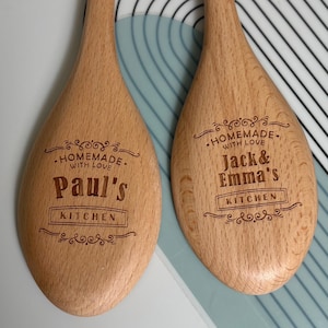 May include: Two light brown wooden spoons with engraved text. The left spoon reads "HOMEMADE WITH LOVE Paul's KITCHEN" with decorative flourishes. The right spoon reads "HOMEMADE WITH LOVE Jack & Emma's KITCHEN" with similar decorative flourishes.