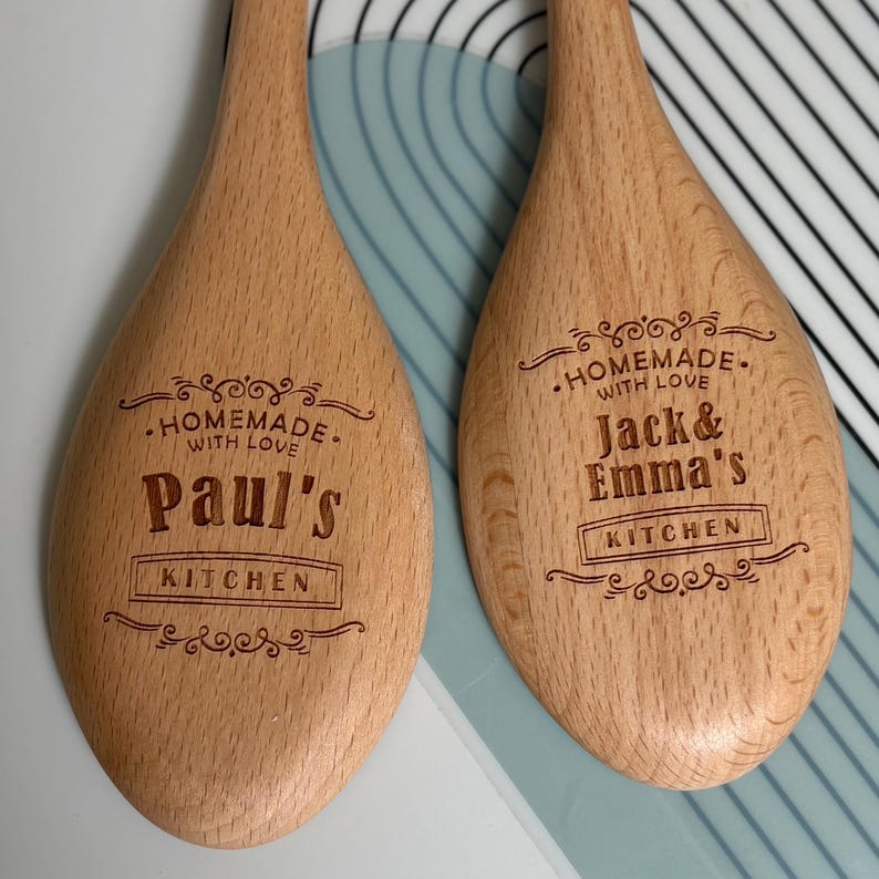 May include: Two light brown wooden spoons with engraved text. The left spoon reads "HOMEMADE WITH LOVE Paul's KITCHEN" with decorative flourishes. The right spoon reads "HOMEMADE WITH LOVE Jack & Emma's KITCHEN" with similar decorative flourishes.