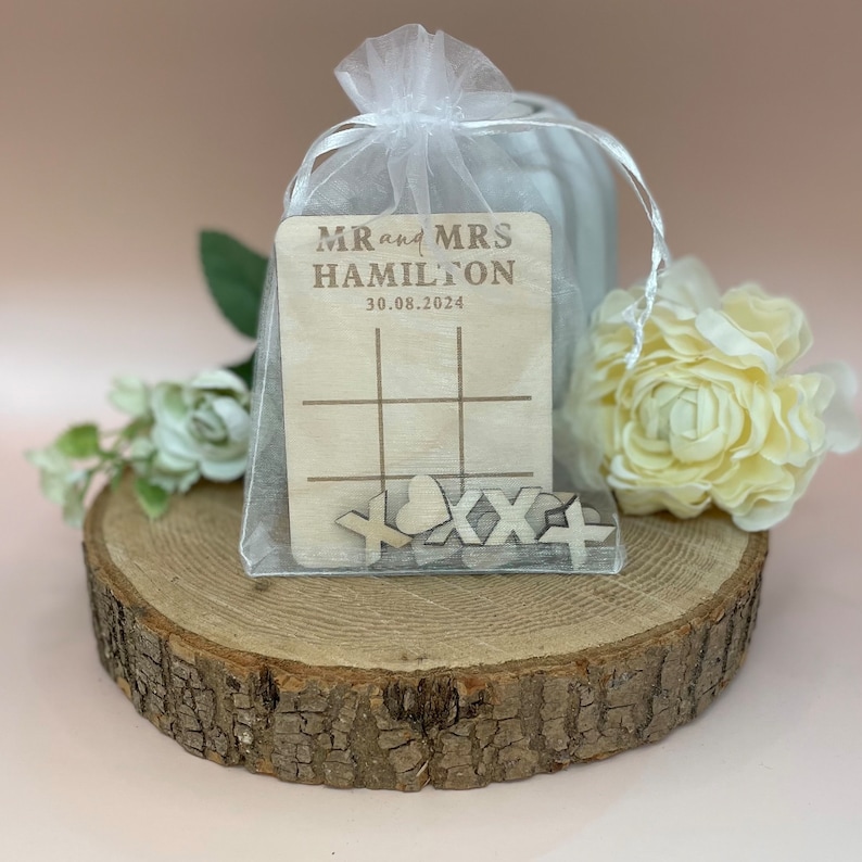 K&ouml;nnte beinhalten: Ein Tic-Tac-Toe-Spiel aus Holz in einem wei&szlig;en Organzas&auml;ckchen. Das Spielbrett ist mit "MR and MRS HAMILTON 30.08.2024" personalisiert. Die Spielsteine sind aus Holz und in Form von Herzen, X und O.