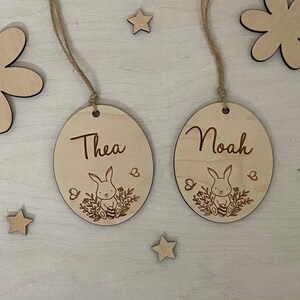 May include: Two wooden Easter egg ornaments with bunny and floral designs. The ornaments are personalized with the names "Thea" and "Noah".