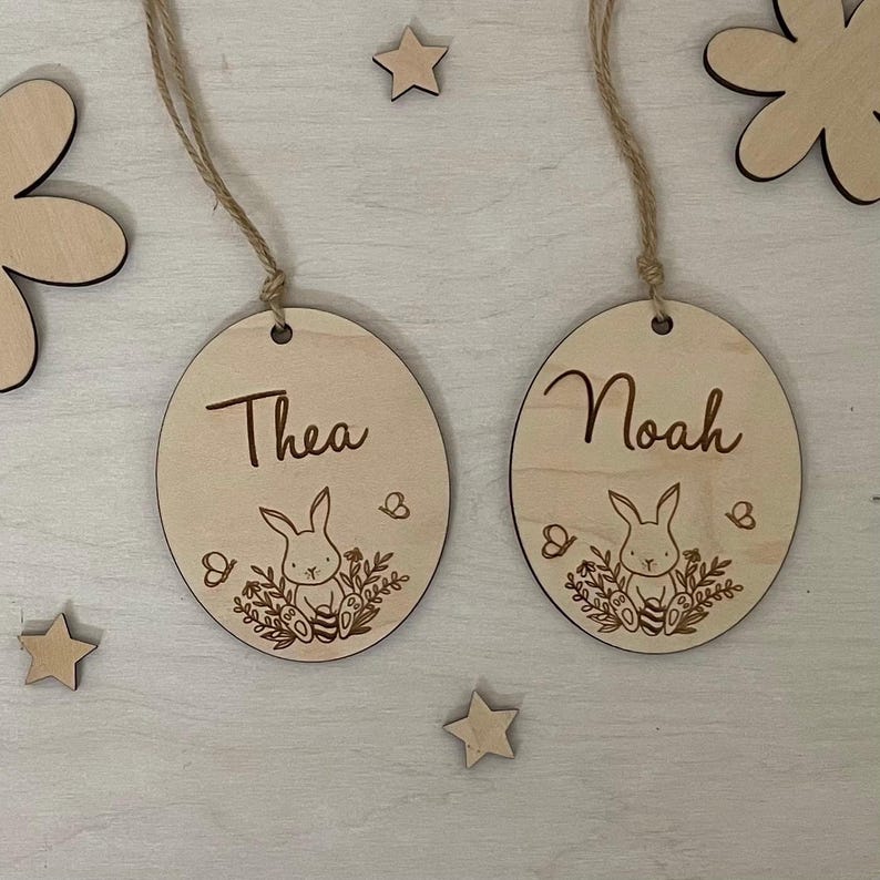 May include: Two wooden Easter egg ornaments with bunny and floral designs. The ornaments are personalized with the names "Thea" and "Noah".