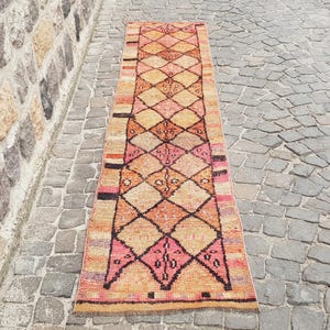May include: A long, narrow runner rug featuring a repeating diamond pattern in shades of orange, pink, and tan. The rug has dark brown geometric outlines and horizontal striped borders at the ends with accents of black, red, and purple throughout the weave.