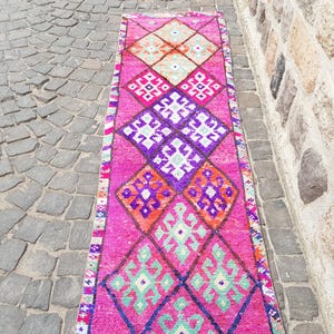 May include: A vibrant, rectangular runner rug with a repeating geometric pattern in shades of pink, purple, orange, and green. The rug is placed on a cobblestone surface, with a stone wall in the background.