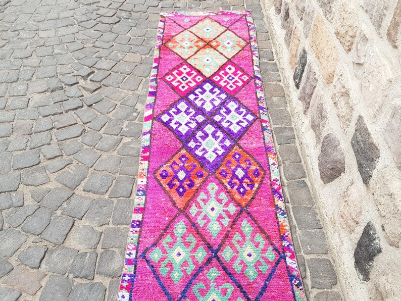 May include: A vibrant, rectangular runner rug with a repeating geometric pattern in shades of pink, purple, orange, and green. The rug is placed on a cobblestone surface, with a stone wall in the background.