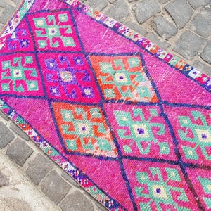 May include: A vibrant, rectangular rug with a repeating diamond pattern. The rug features a bright pink background with geometric shapes in teal, orange, and purple. The edges are trimmed with a patterned border.