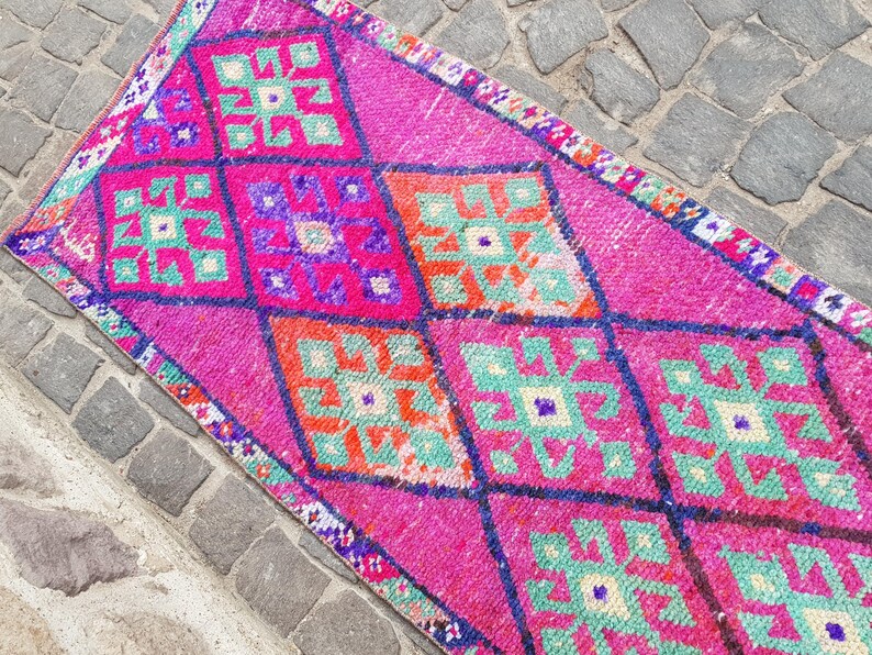 May include: A vibrant, rectangular rug with a repeating diamond pattern. The rug features a bright pink background with geometric shapes in teal, orange, and purple. The edges are trimmed with a patterned border.