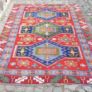 Könnte beinhalten: Ein rechteckiger Teppich mit einem leuchtend roten Hintergrund und drei großen blauen rautenförmigen Medaillons. Das Design umfasst geometrische Muster in Grün, Orange und Weiß sowie eine breite Bordüre mit sich wiederholenden Motiven.