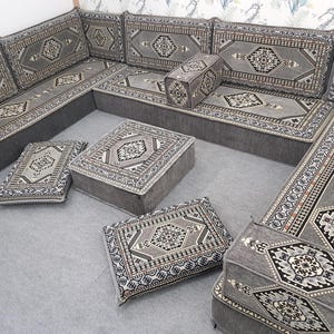 May include: A gray sectional sofa with matching patterned cushions and ottomans. The cushions and ottomans feature a detailed geometric design in shades of gray, black, and beige. The sofa is arranged in an L-shape, with several square cushions and a rectangular ottoman.