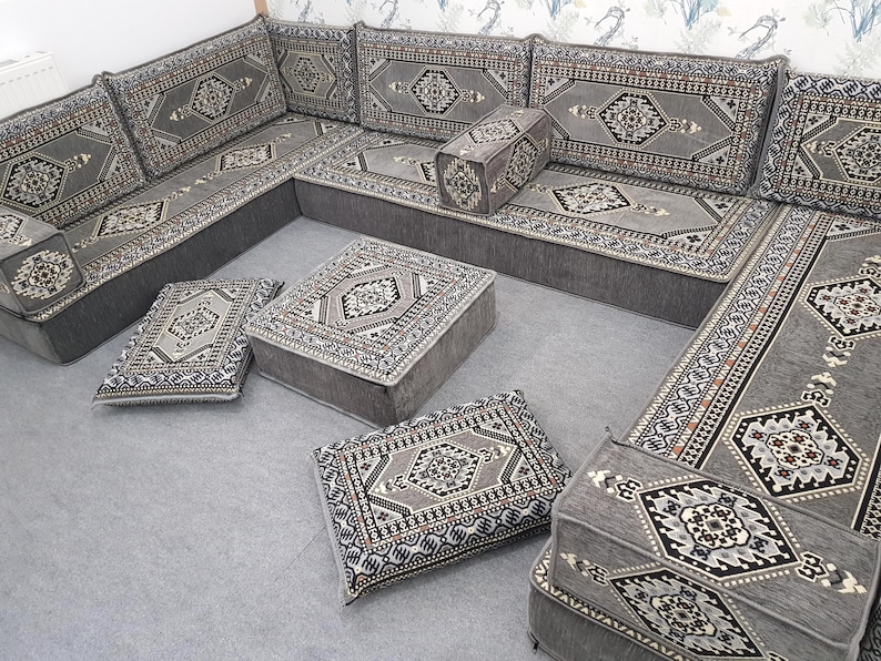 May include: A gray sectional sofa with matching patterned cushions and ottomans. The cushions and ottomans feature a detailed geometric design in shades of gray, black, and beige. The sofa is arranged in an L-shape, with several square cushions and a rectangular ottoman.
