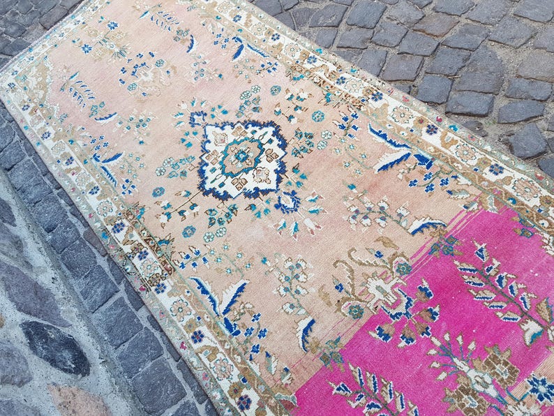 May include: A rectangular area rug featuring a faded peach background with a central dark blue and white floral medallion. The design includes intricate botanical patterns in shades of blue, green, and cream, with a vibrant hot pink section at one end.