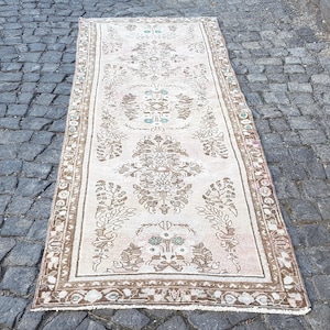 May include: A rectangular runner rug featuring a faded cream background with intricate brown floral and botanical patterns. The design includes a central medallion motif and a detailed border, with subtle accents of muted blue and pink throughout.