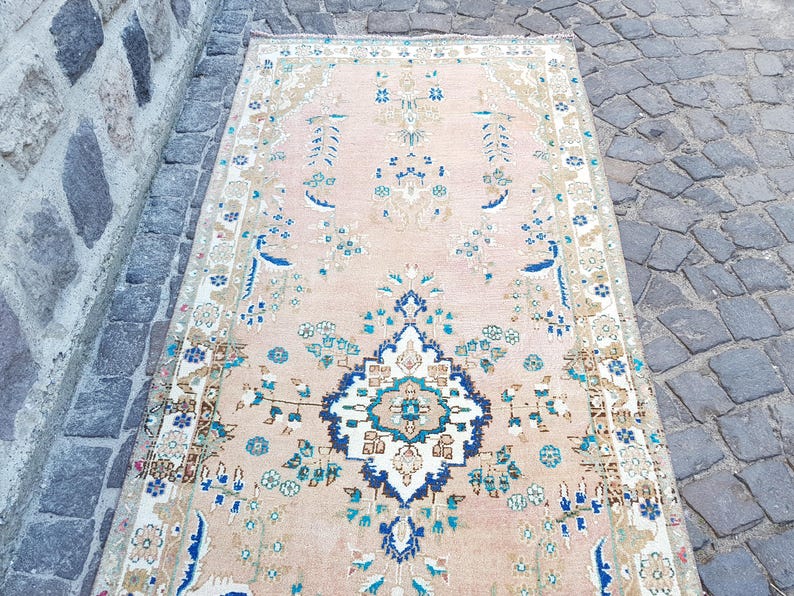 May include: A rectangular runner rug featuring a soft peach background with a central dark blue and white medallion. The rug is decorated with intricate floral patterns in shades of teal, light blue, and beige, framed by a detailed border.
