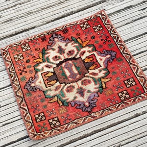 May include: A square rug featuring a vibrant red background with a central cream, brown, and purple geometric medallion pattern. The border displays a repeating dark brown and white zigzag motif, accented by small multi-colored floral squares.