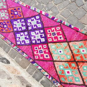 May include: A vibrant, rectangular rug with a repeating geometric pattern. The rug features a bright pink background with diamond shapes in purple, orange, and turquoise. The rug is likely made of wool or a similar material.