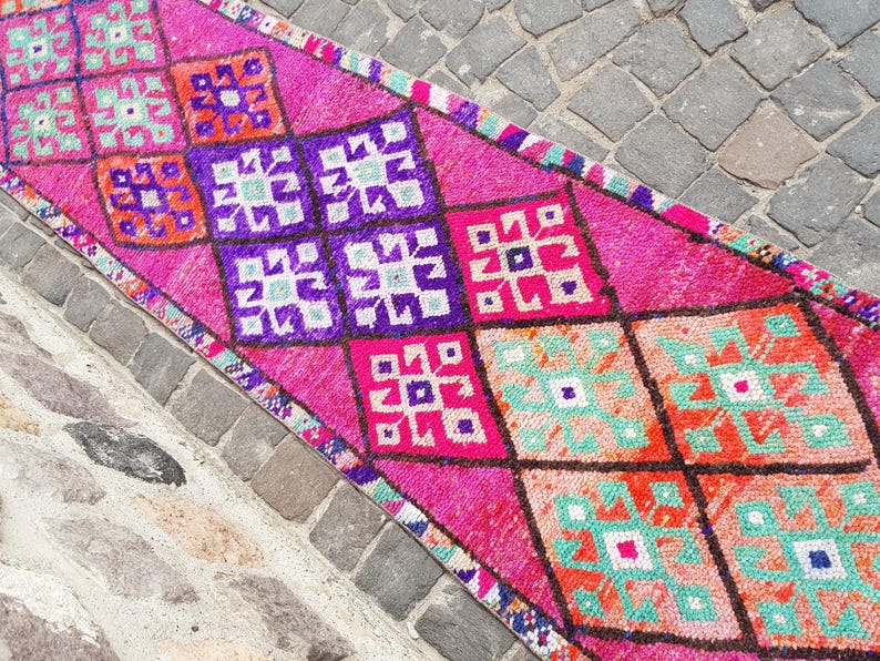 May include: A vibrant, rectangular rug with a repeating geometric pattern. The rug features a bright pink background with diamond shapes in purple, orange, and turquoise. The rug is likely made of wool or a similar material.