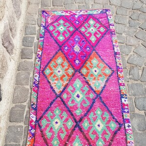 May include: A vibrant, rectangular rug with a pink background and geometric diamond patterns in orange, green, and purple. The rug's border features a detailed, colorful design. This rug is suitable for home decor.