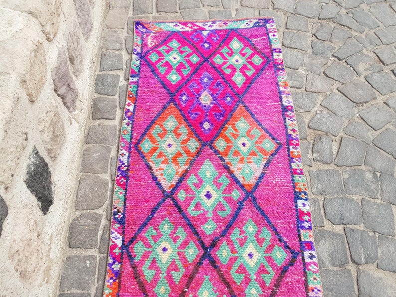 May include: A vibrant, rectangular rug with a pink background and geometric diamond patterns in orange, green, and purple. The rug's border features a detailed, colorful design. This rug is suitable for home decor.