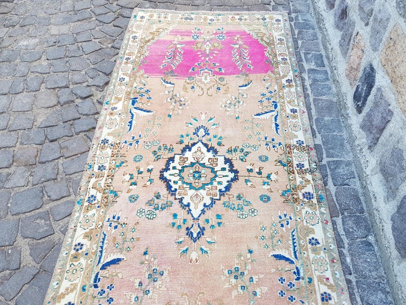 May include: A rectangular runner rug featuring a central dark blue and white geometric medallion on a light peach background. The rug includes a vibrant pink section at one end, decorated with intricate floral patterns in blue, green, and white.
